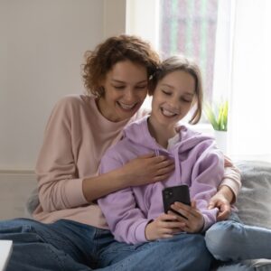 Happy teen daughter and young mum using smartphone at home taking selfie