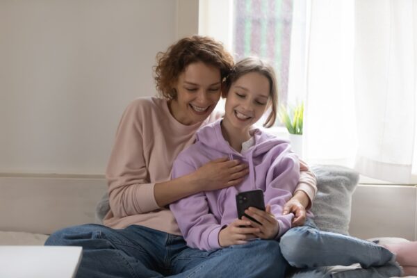 Happy teen daughter and young mum using smartphone at home taking selfie